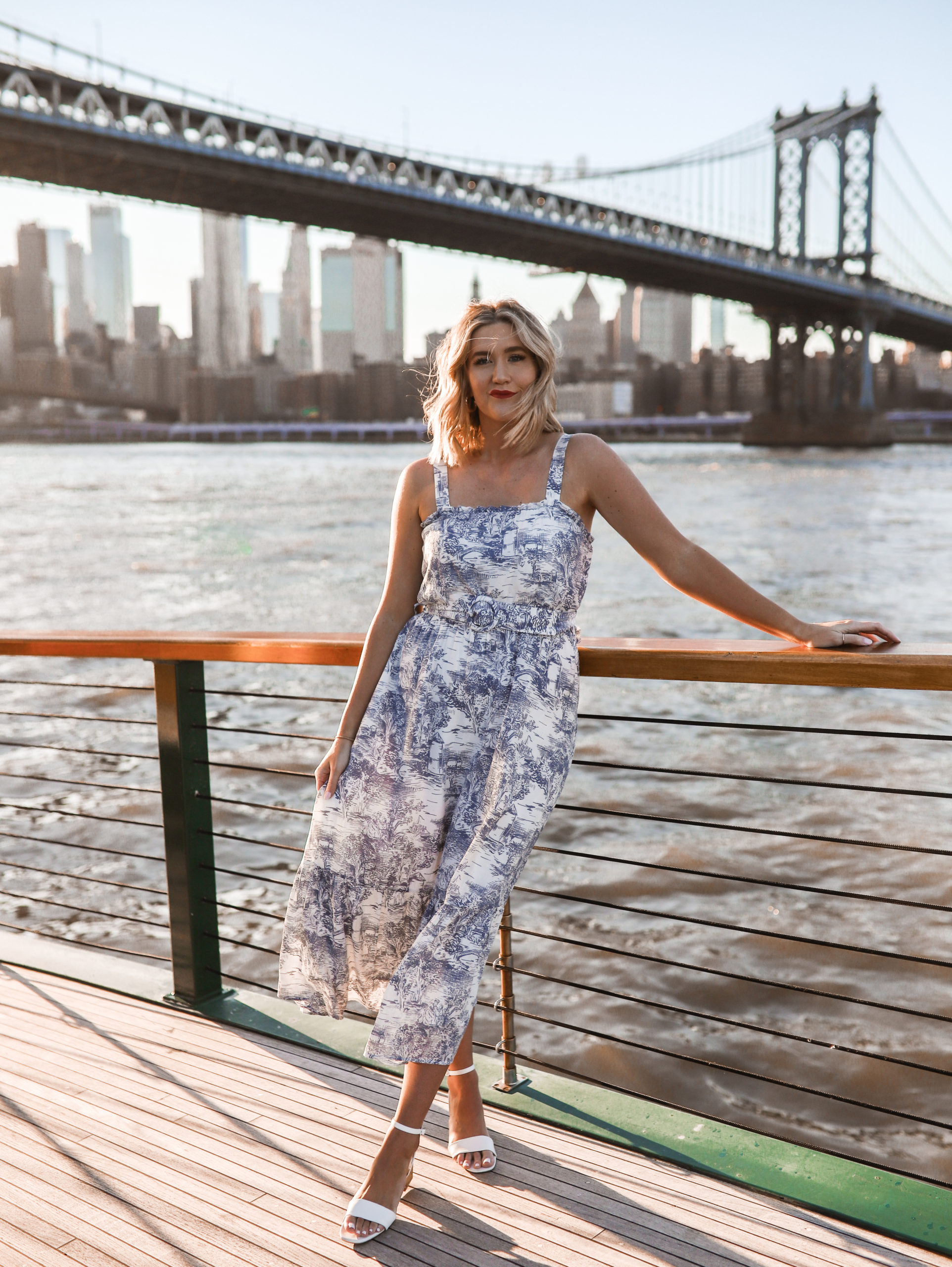 Anna standing on a pier in Brooklyn with the Brooklyn Bridge behind her as she's wearing a dress.