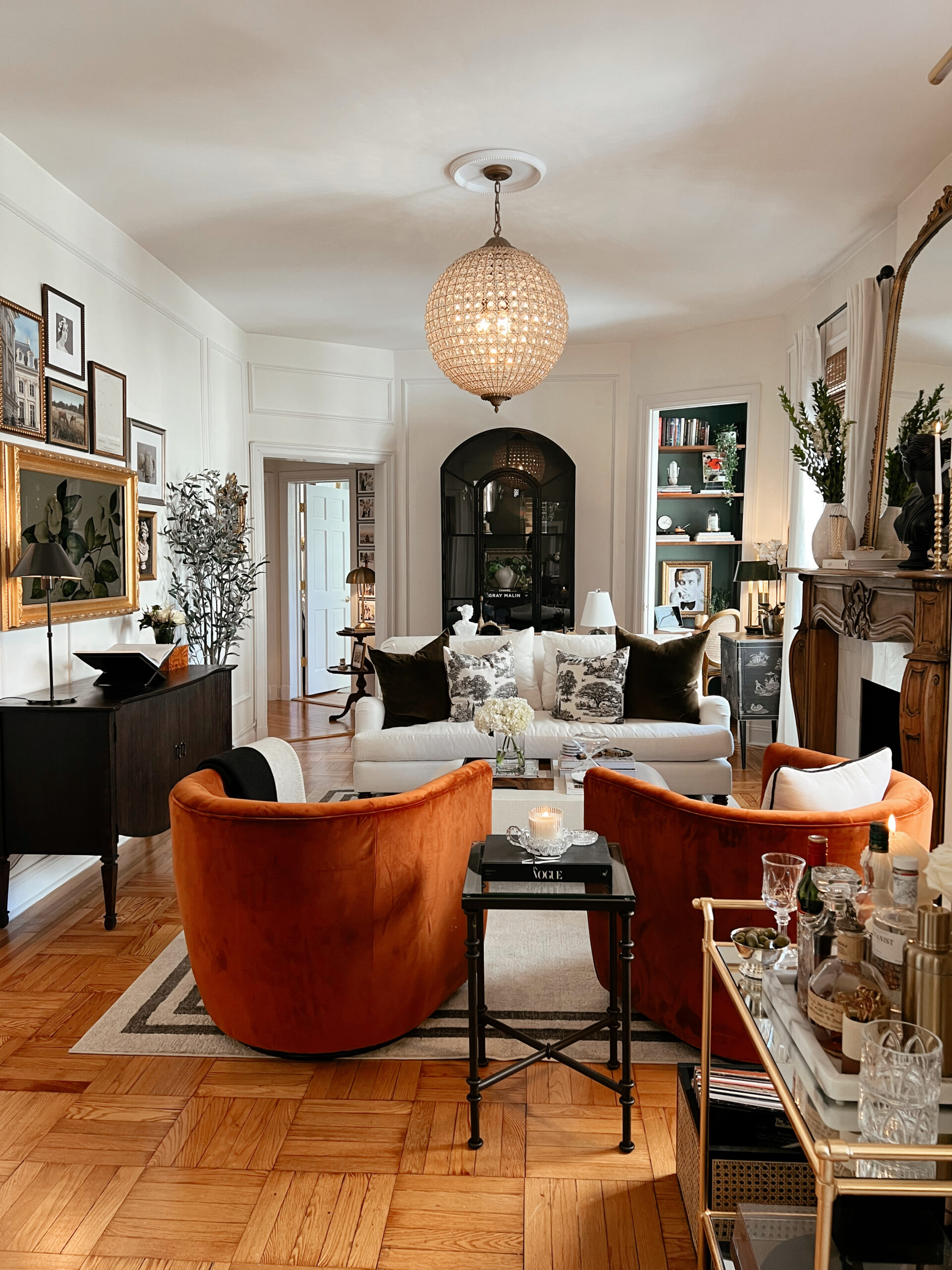 Anna's living room with two large orange chairs, a white couch with dark colored pillows, and a gorgeous chandelier in frame.
