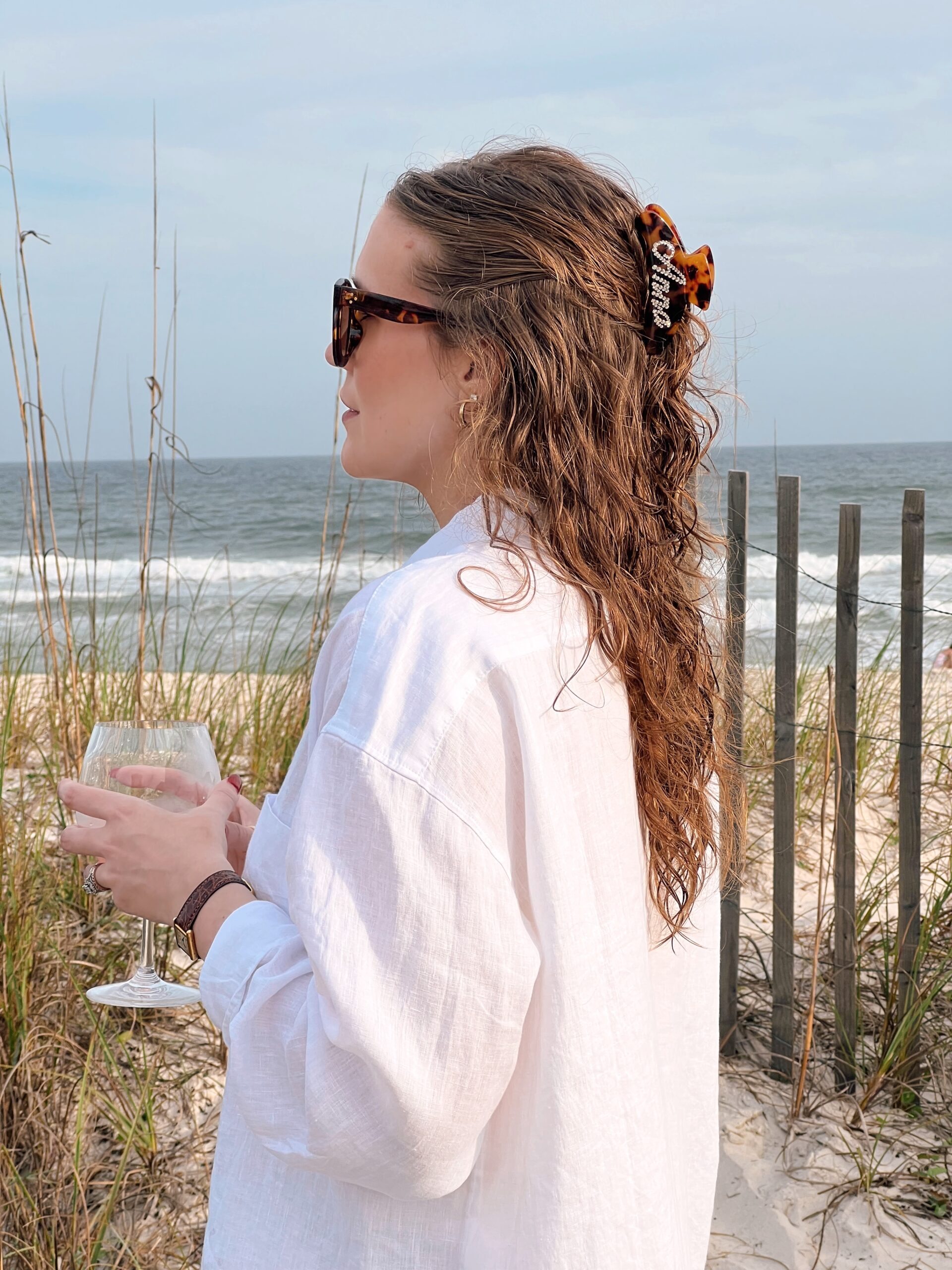 Anna standing on the beach with a glass of wine and sunglasses on.