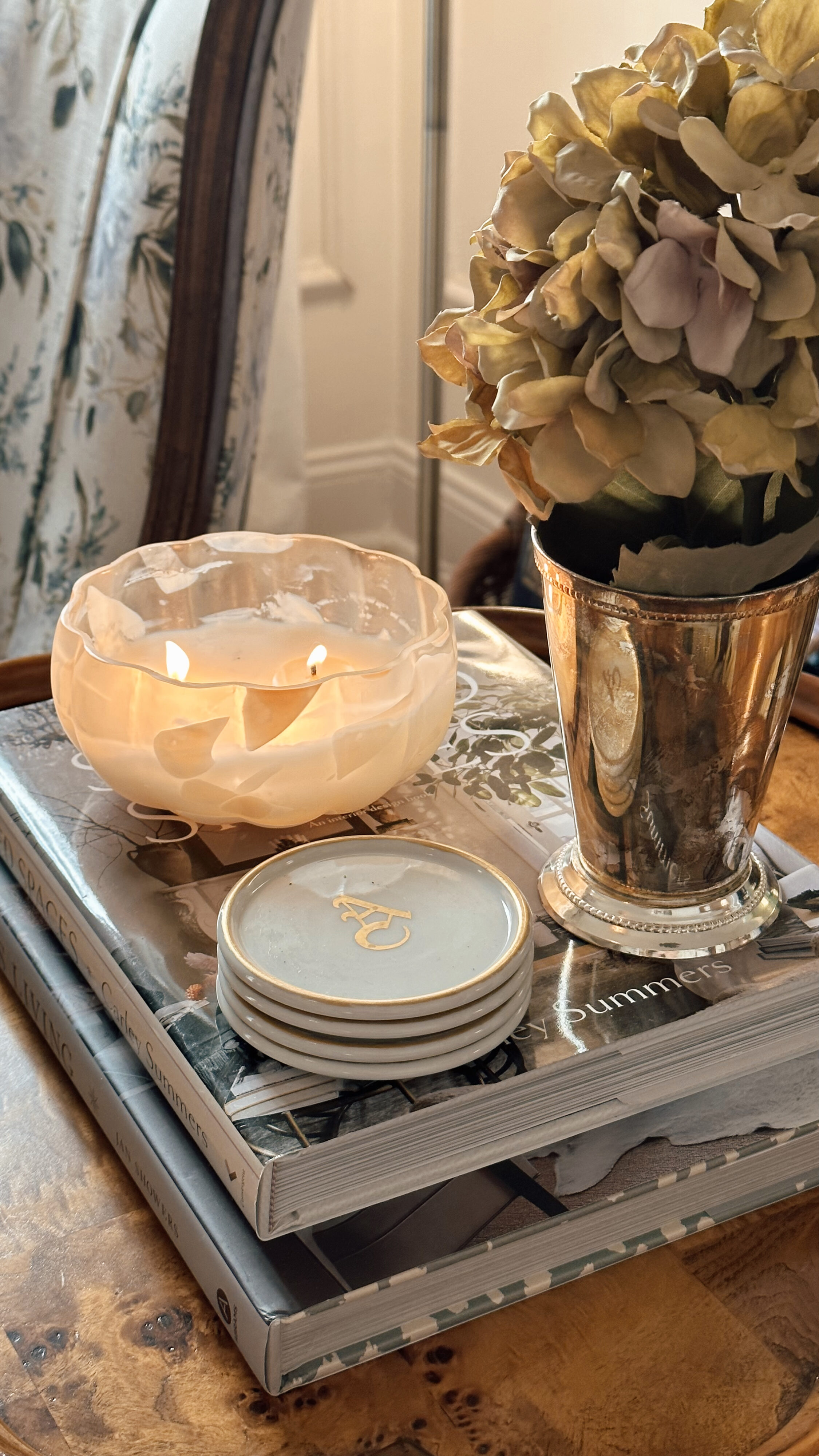 A cute section on a table with coffee table books, a candle, coasters, and a floral arrangement.