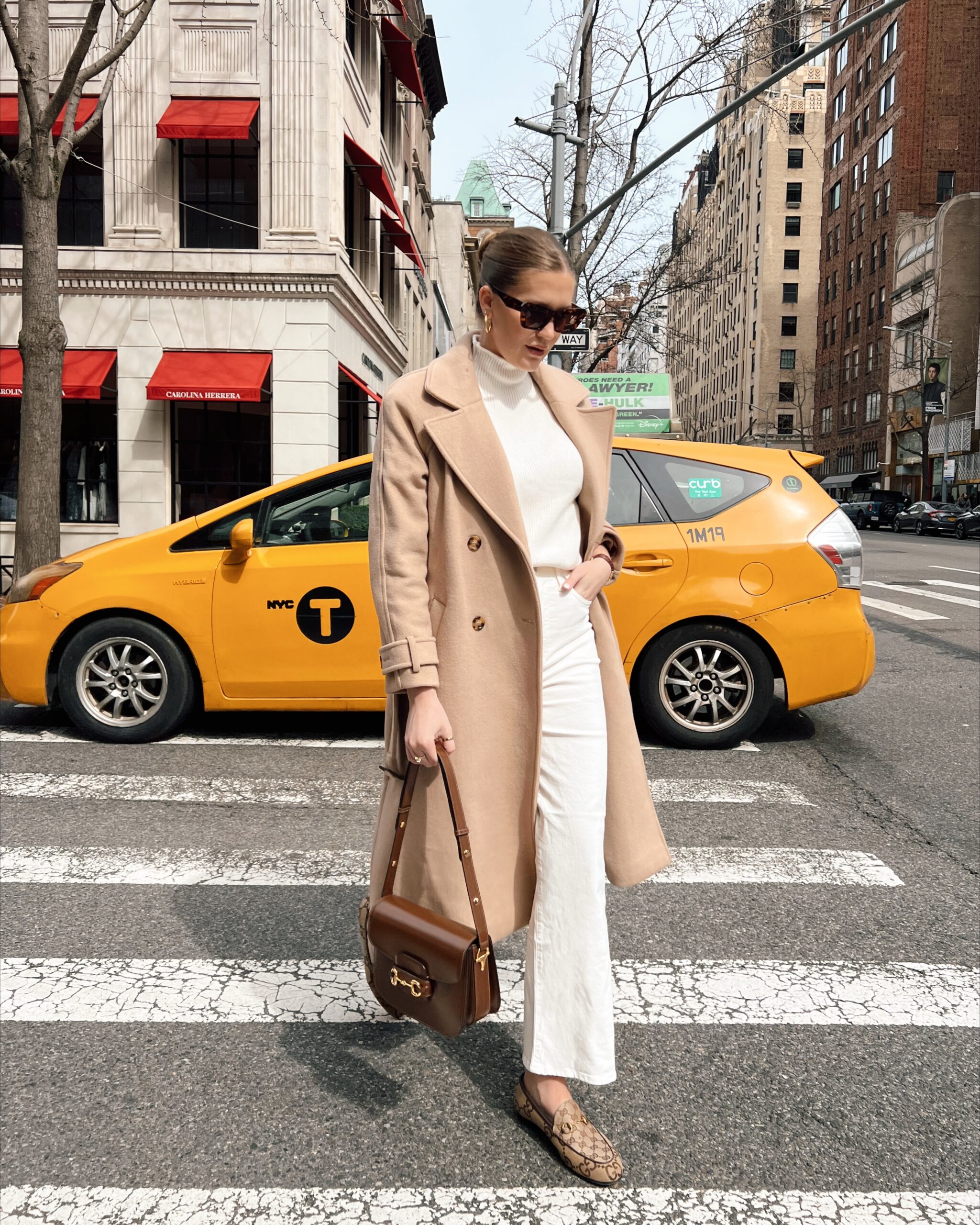 Anna walking in a trench coat in front of a yellow taxi in New York City.