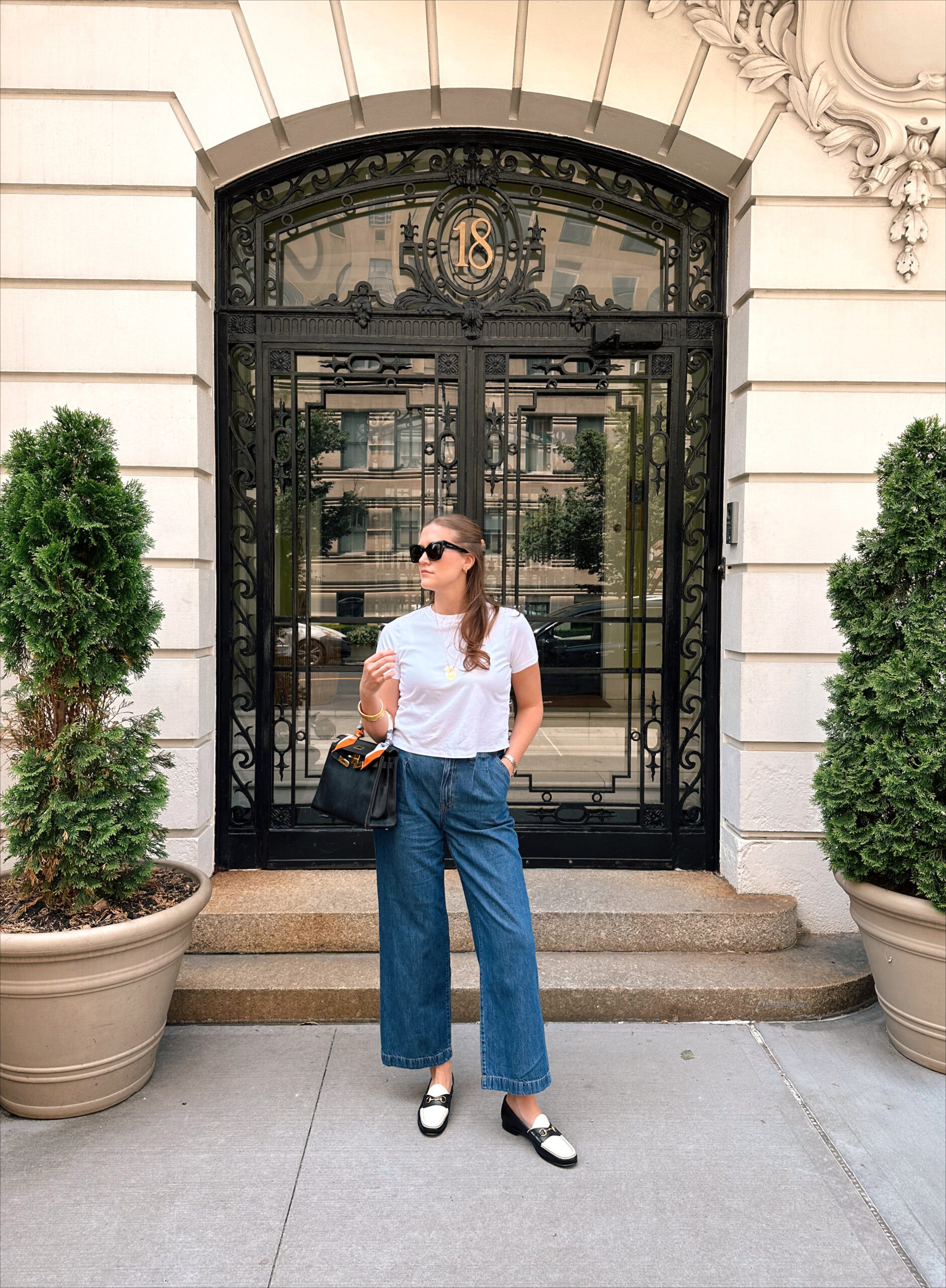 Anna standing in front of a beautifully ornate door in New York City wearing a white top and blue jeans.